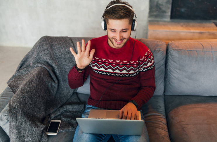 young smiling man sitting home winter having conversation online waving hand saying hello wearing red sweater working laptop freelancer listening headphones studying online